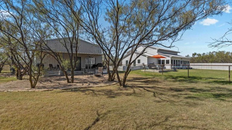 A fenced yard with two light-colored metal buildings, trees providing shade, a patio area with outdoor seating, and a large grassy lawn under a blue sky.