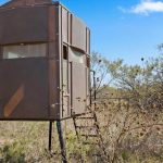 A metal hunting blind raised on four legs stands in a dry, grassy area surrounded by shrubs and small trees under a blue sky with scattered clouds. A small staircase leads up to its door.