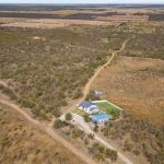 Aerial view of a rural property with a house, outbuildings, a pond, and surrounding dry, brushy landscape. Dirt roads and open fields stretch into the distance under a cloudy sky.