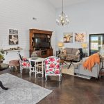 Bright, spacious living room with a farmhouse style. Features gray sofas, floral armchairs, a chandelier, TV, vintage decor, and large windows with a view. White shiplap walls and rustic wooden furniture add charm.