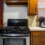 A kitchen with a black and stainless steel gas stove, wooden cabinets, a countertop with a coffee maker and another appliance, a window with wooden blinds, and a door with a small pet door at the bottom.