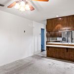 A room with white walls and carpet, a ceiling fan with lights, and a wooden kitchen counter with cabinets dividing the space from the kitchen area, which contains appliances and a window.