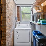 A small laundry room with a white dryer, shelves holding various items, a recycling bin, and a trash bin, featuring exposed brick and wood paneling. A window at the end lets in natural light.