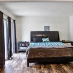 A bedroom with a large bed covered in a blue-patterned blanket, two nightstands on either side, dark curtains over a glass door, and wood-patterned flooring. Walls and furnishings are neutral-toned.