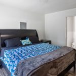 A bedroom with a large black bed, blue geometric-patterned blanket, black pillows, two mirrored nightstands, and a doorway leading to a bathroom with visible toilet. The room has white walls and dark wood flooring.