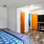 A bedroom with wood-look flooring, a bed with blue-patterned bedding, a dark dresser, a large TV, and three open doors—one leading to a bathroom, another to a hallway, and a third to a blue-walled room.