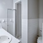 A small bathroom with white tiled walls, a sink with a faucet, a mirror, a toilet, and a frosted glass shower door under a wall-mounted light.