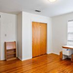 A room with wooden floors, white walls, a double closet with wooden doors, a window with blinds, a desk, a cat tree, a small shelf, and an open door leading to a hallway.