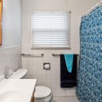 A bathroom with white tile walls, a white sink and toilet, a window with closed blinds, and a shower covered by a blue patterned curtain. A towel rack holds a teal and a black towel.