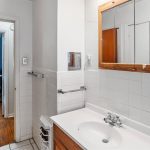 A bathroom with white tiled walls, a sink with a wooden cabinet, a mirrored medicine cabinet, and a towel rack. An open door leads to a room with wood floors and a lamp visible in the background.
