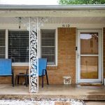 A small brick house front porch with two blue chairs, a small table, and decorative white metal post. A glass door is next to a window with white blinds. Pebbles line the ground near the porch. House number 5112 is above the door.