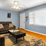 A cozy living room with blue walls, hardwood floors, a ceiling fan, gray sofas with patterned pillows, a black coffee table, a rug, lamps, and large windows with white blinds letting in natural light.