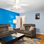 A living room with two gray sofas, a black coffee table, a lamp, and a colorful rug on a wooden floor. The walls are painted blue and light blue, and there is a ceiling fan with lights.
