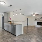 Open-concept kitchen and living area with gray cabinets, granite counters, hanging lights, tile flooring, built-in shelving, fireplace, and large windows allowing natural light.