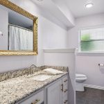 A bathroom with a granite countertop, gold-framed mirror, potted orchid, white cabinets, a toilet, window with blinds, and a shower with a white curtain. The walls are light, and the floor is gray tile.
