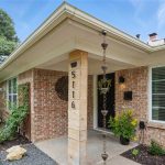 Single-story brick house with a covered front porch, a rain chain, plants along the walkway, and large windows with white blinds. The address “5116” is displayed vertically on a porch post.
