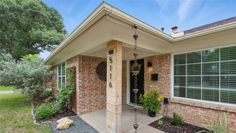 Single-story brick house with a covered front porch, a rain chain, plants along the walkway, and large windows with white blinds. The address “5116” is displayed vertically on a porch post.