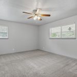 A spacious, empty room with light gray walls, carpeted floor, two small windows on adjacent walls, and a ceiling fan with lights in the center of the ceiling.