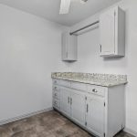 A small laundry or utility room with gray cabinets, a granite countertop, overhead storage, a ceiling fan, and a door with a window. The walls are painted white and the flooring is dark vinyl or tile.