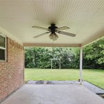 Covered patio with a ceiling fan, concrete floor, and brick wall on the left. The patio overlooks a grassy backyard with trees, a swing set, and a gravel border along the edge.