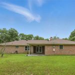 A single-story brick house with a covered back patio and large grassy backyard, surrounded by a wooden fence and mature trees under a clear blue sky.