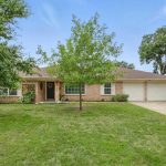 Single-story brick house with a brown roof, green lawn, two large trees in the front yard, two-car garage, and a paved driveway on a sunny day.