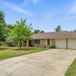 Single-story brick house with a two-car garage, wide driveway, and a grassy front yard with trees. The house is on a corner lot in a suburban neighborhood under a blue sky with light clouds.