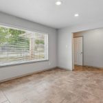 Empty room with tiled floor, large window with blinds letting in natural light, light gray walls, recessed ceiling lights, and a doorway leading to another room with a closed white door.