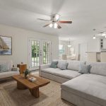 A bright living room with a large gray sectional, modern wooden coffee table, armchair, and art of a sailboat. The space opens to a dining area and kitchen with bar stools and pendant lights. French doors lead outside.