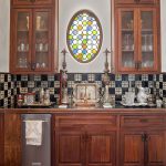 A kitchen wet bar with dark wood cabinets, glass doors, a small sink, checkered tile backsplash, and two ornate lamps. Above the sink is an oval stained glass window with colorful circular patterns.