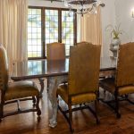 A dining room with a dark wooden table, six gold-upholstered chairs, hardwood floors, cream-colored curtains, a chandelier, a large vase with flowers, and a wooden door in the background.
