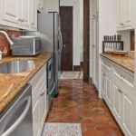 A narrow kitchen with white cabinets, brown granite countertops, terracotta tile floor, stainless steel appliances, a microwave, knife set, and a silver sink. The backsplash is red tile, and a rug lies on the floor.