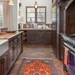 A rustic kitchen with dark wood cabinets, granite countertops, a farmhouse sink, colorful tiled backsplash, stainless steel appliances, and a vibrant red patterned rug on a herringbone tile floor.