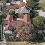 Aerial view of a suburban neighborhood with houses featuring red-tiled roofs, trees lining the streets, a roundabout with a landscaped centerpiece, and a mix of lawns and driveways.