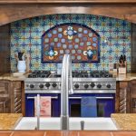 A kitchen with a colorful tiled backsplash, dark wooden cabinets, a blue stove, granite countertops, knives, utensils, and a sink with a silver faucet in the foreground.