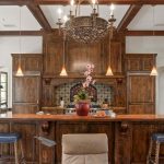 A rustic kitchen with dark wood cabinets, a large island with rope-accented stools, a decorative tile backsplash, and a chandelier above. A potted orchid sits on the island, and arched windows and doorways are visible.