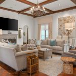A spacious living room with terracotta tile flooring, beige sofas, a recliner, a wall-mounted TV above a fireplace, large windows with curtains, and decorative ceiling beams and chandelier.