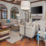 A spacious living room with large arched windows, light-colored sofas, a fireplace, a TV above the mantel, a dining table with chairs, and a floor lamp beside stacked books on the terracotta-tiled floor.