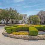 A roundabout with neatly trimmed hedges and colorful flowers in the center, surrounded by upscale houses with varied architecture under a blue sky.