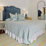 A cozy bedroom with a blue upholstered headboard, pastel bedding, two floral pillows, mirrored nightstand, tall mirror, wicker chair, and striped rug on terracotta tile floor. A bathroom is visible in the background.