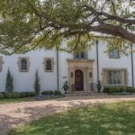 A large, white, two-story house with arched windows and a wooden front door, surrounded by neatly trimmed bushes and a curved stone driveway under a large, leafy tree.