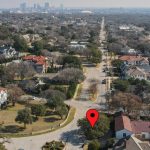 Aerial view of a suburban neighborhood with tree-lined streets and large houses; a red location pin marks a spot near a street corner, with a city skyline visible in the background.