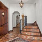 Elegant entryway with decorative tiled staircase, wrought iron railing, arched doorways, wooden floors, and ornate light fixtures. A hallway leads to other rooms, and a glimpse of a kitchen is visible to the left.
