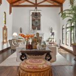 A spacious, elegant living room with high wooden ceilings, large arched windows, a tapestry on one wall, ornate furniture, a large potted plant, and a central table with flowers and decorative items.