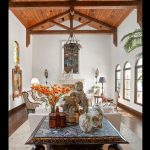 A spacious living room with wooden beams, arched windows, elegant sofas, a chandelier, a marble table with vases and flowers, and a white fireplace against white walls. Ornate decor and artwork complete the classic look.