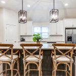 Modern kitchen with white cabinets, stainless steel appliances, a large island with three wooden bar stools, pendant lights above, and a potted plant in the corner. A window lets in natural light over the sink.