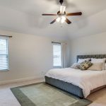 A spacious bedroom with a large bed, white bedding, and decorative pillows. There are two windows with beige curtains, a wooden nightstand, a dresser, a ceiling fan with lights, and a green area rug on the floor.
