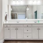 Modern bathroom with double sinks, white cabinets, a large wall mirror, two silver towel racks with gray towels, and bright overhead lighting. The space is clean and uncluttered.