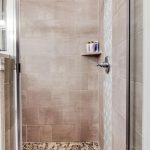 A walk-in shower with beige tiled walls, a built-in corner shelf holding toiletry bottles, a metal shower handle, and a pebble stone floor. A window with blinds is partially visible on the left.