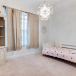 A bright, tidy child's bedroom with a white bed covered in a pink polka-dot blanket, a plush carpet, beige curtains over a window, a bookshelf with toys and books, and a decorative chandelier hanging from the ceiling.